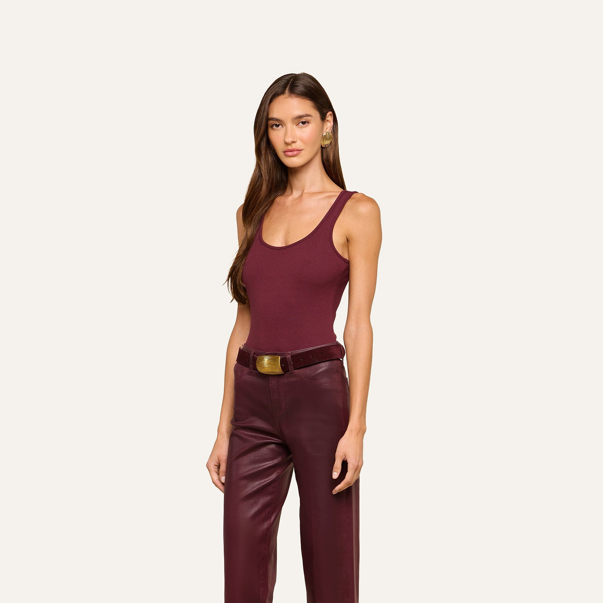 A woman with long brown hair wears a sleeveless burgundy Nica Scoopneck Bodysuit paired with matching leather pants and a brown belt, standing against a plain off-white background.