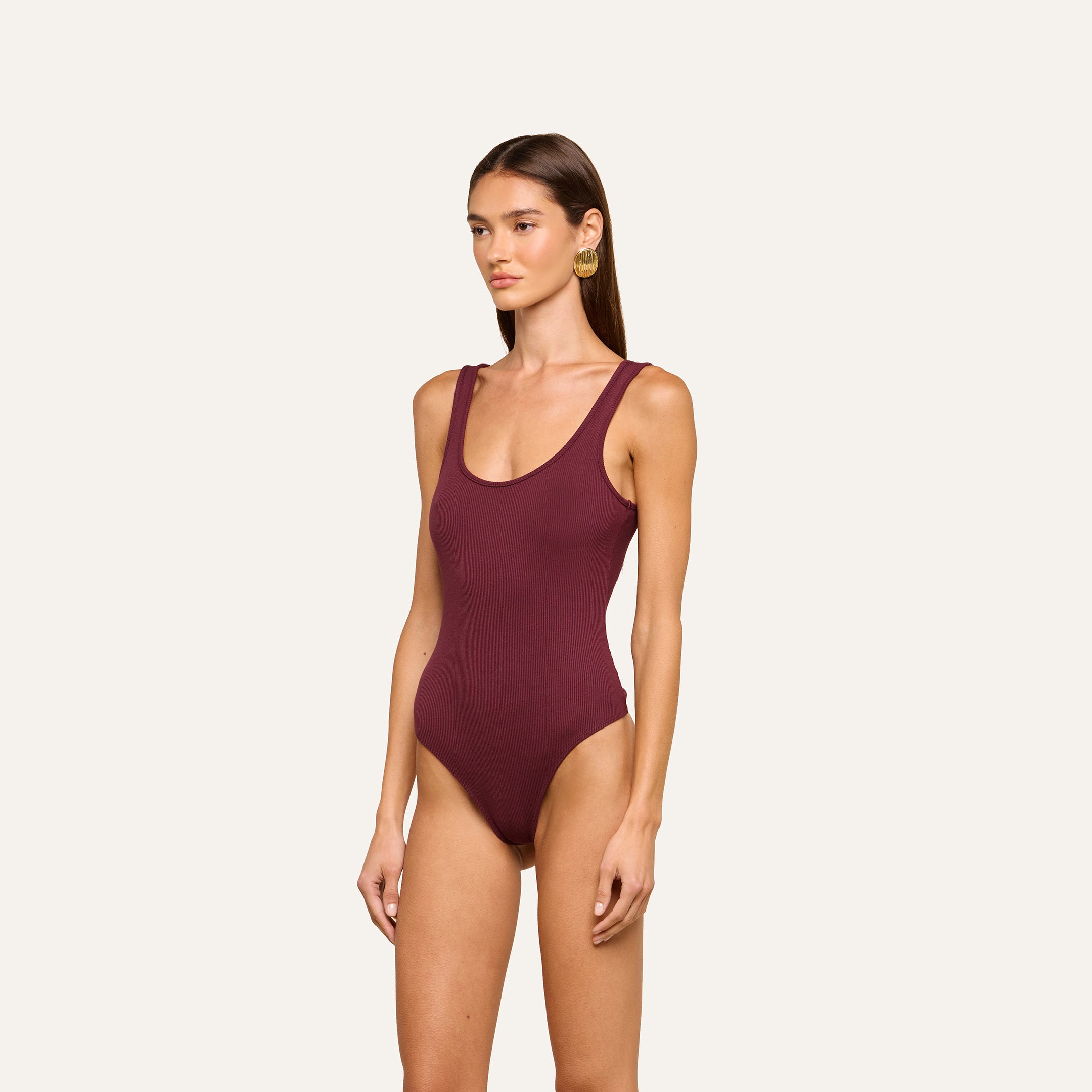 A woman stands against a plain background wearing the Nica Scoopneck Bodysuit, a sleeveless base layer in dark maroon with a scoop neckline, high-cut legs, and high-recovery stretch fabric. She looks slightly to her left.