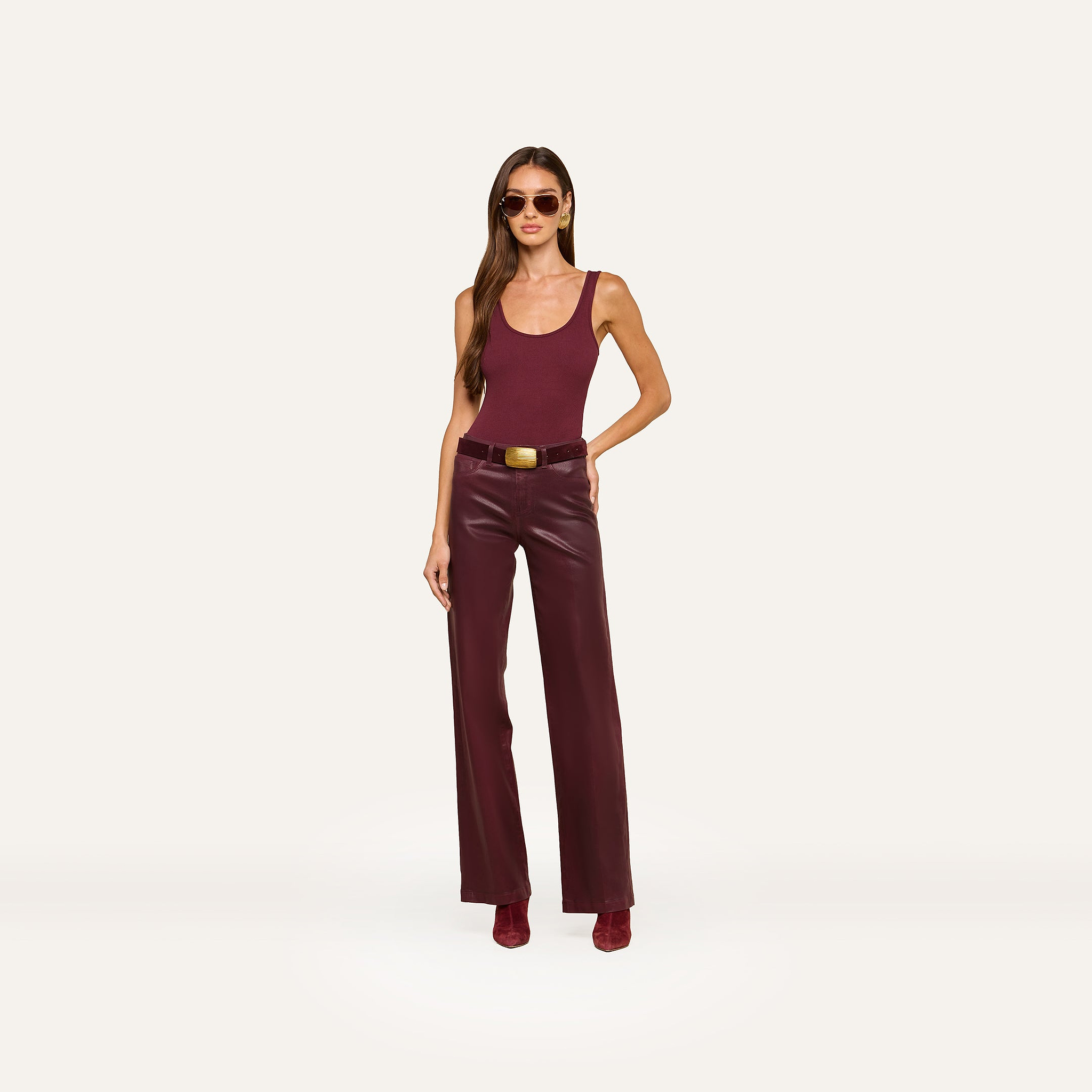 A woman wearing sunglasses, a burgundy Nica Scoopneck Bodysuit styled as a tank top, high-waisted burgundy leather pants with high recovery stretch, and matching boots stands against a plain white background.