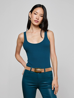 A woman with long dark hair models the L'AGENCE Nica Scoopneck Bodysuit in teal, paired with teal pants, a tan belt with a large buckle, and gold hoop earrings. She stands against a plain gray background, one hand on her hip.