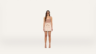 A woman wearing sunglasses, a sleeveless beige top, a light pink skirt with a bow, and heeled sandals stands against a plain white background.