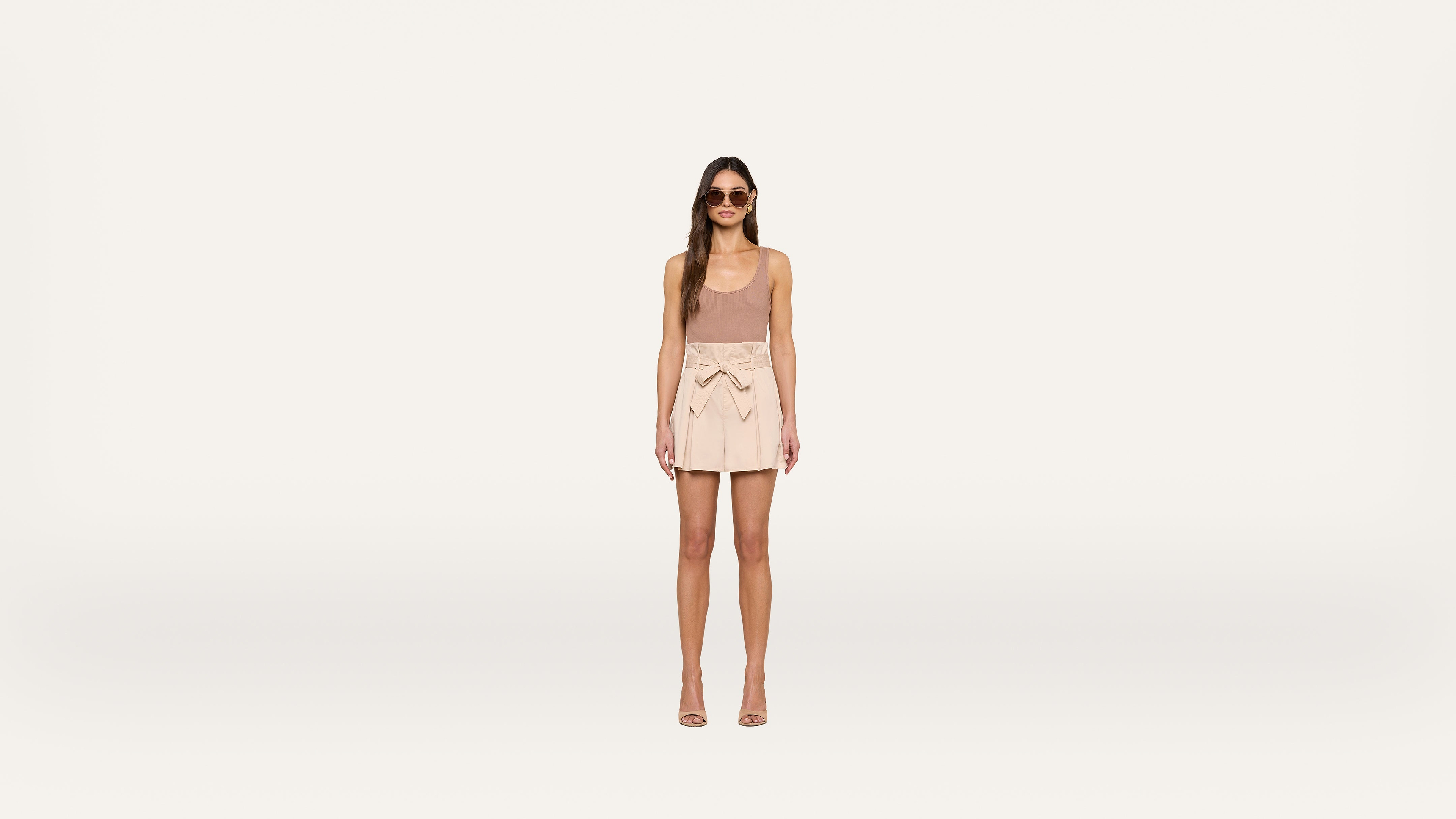 A woman wearing sunglasses, a sleeveless beige top, a light pink skirt with a bow, and heeled sandals stands against a plain white background.