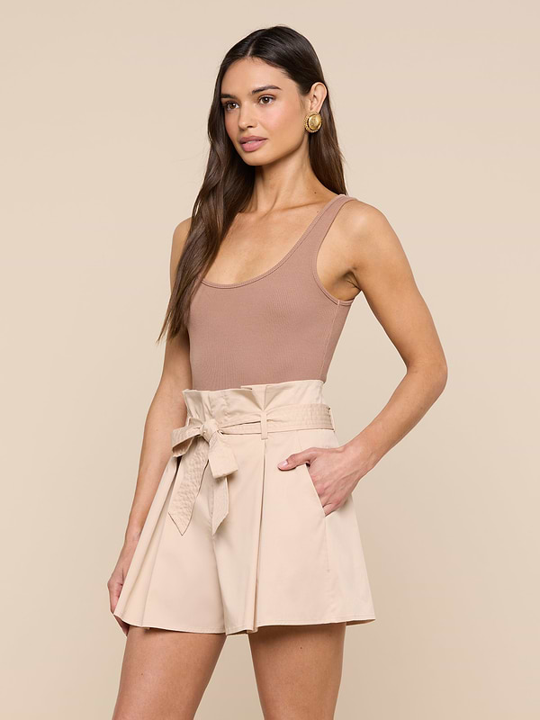 A woman wearing the Nica Scoopneck Bodysuit in sleeveless, scoop-neck beige with high recovery stretch rib and high-waisted, light beige shorts with a tied belt stands with one hand in her pocket against a plain background.