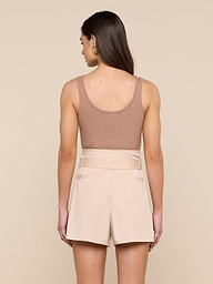 A woman with long brown hair, wearing the Nica Scoopneck Bodysuit—a sleeveless taupe top with a sculpted fit—and high-waisted beige shorts stands facing away from the camera against a plain light background.