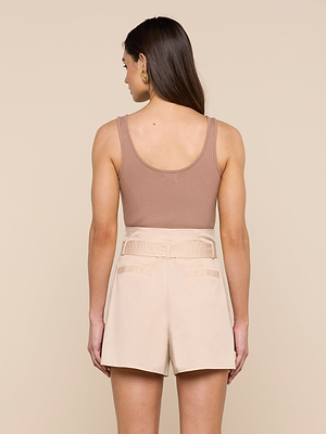 A woman with straight brown hair and gold hoop earrings faces away, wearing the Nica Scoopneck Bodysuit in taupe and high-waisted light beige shorts with pockets. The background is beige.