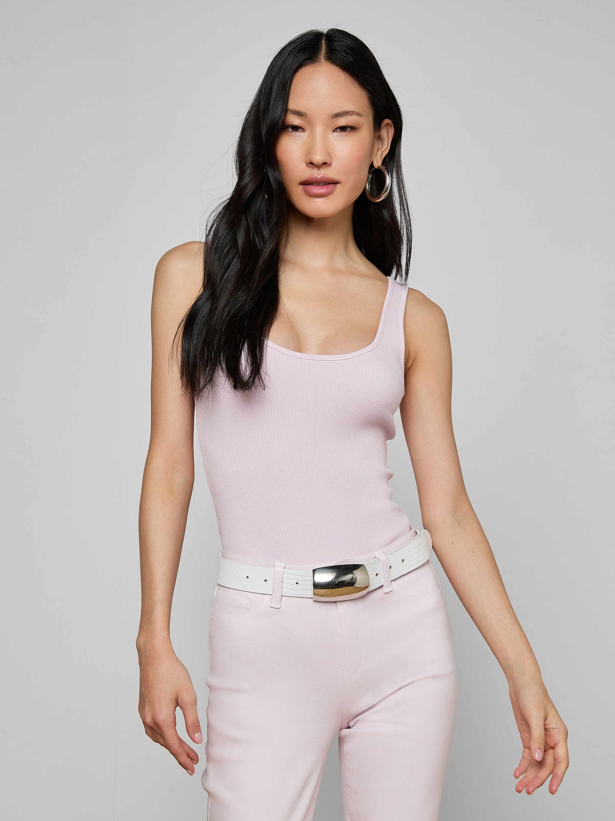 A woman in the Nica Scoopneck Bodysuit in light pink, paired with matching pink pants, a white belt with a silver buckle, and hoop earrings stands against an off-white background.