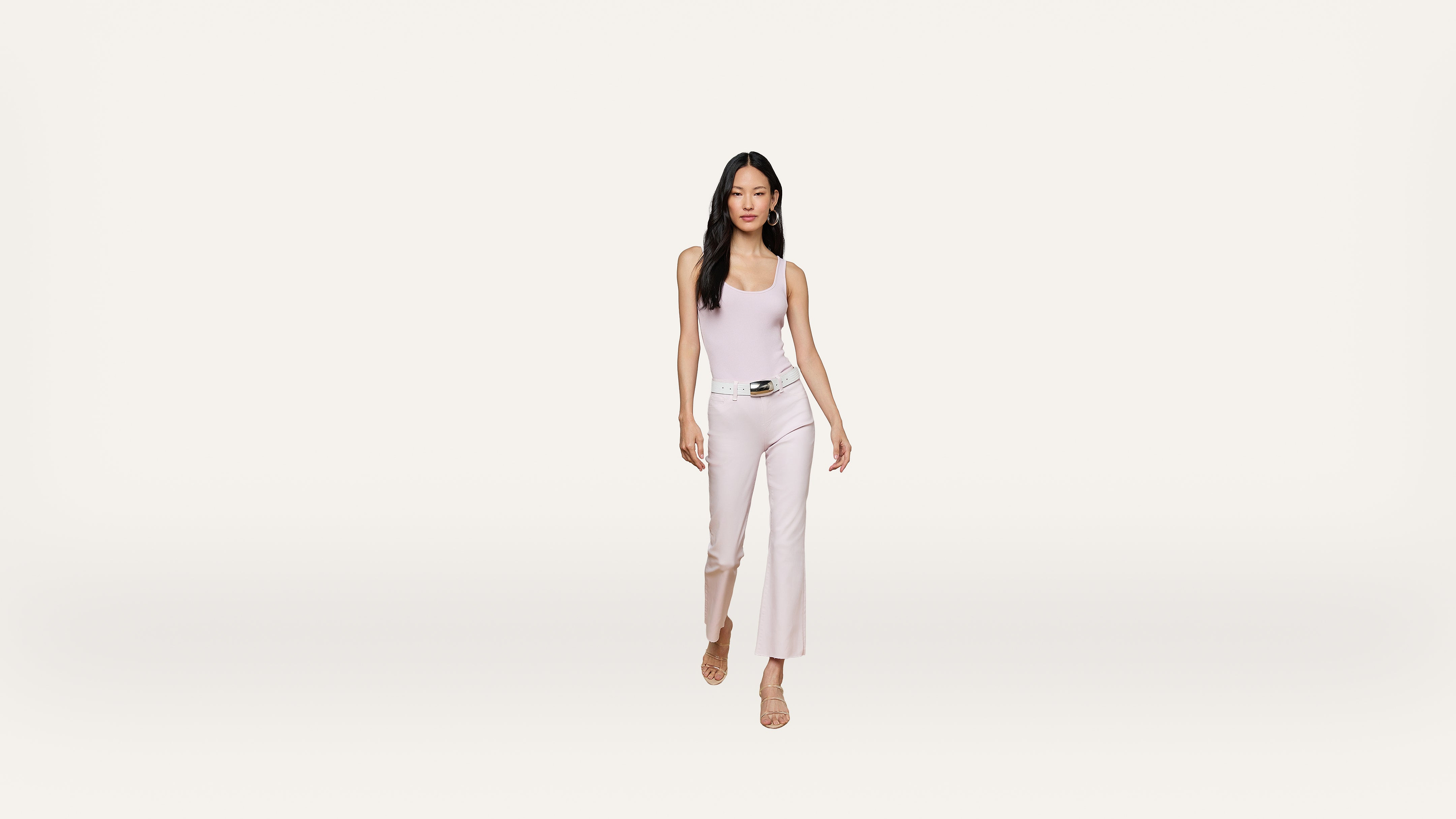 Woman with long dark hair wears a light pink sleeveless top, white pants with a belt, and beige sandals, walking forward against a plain white background.