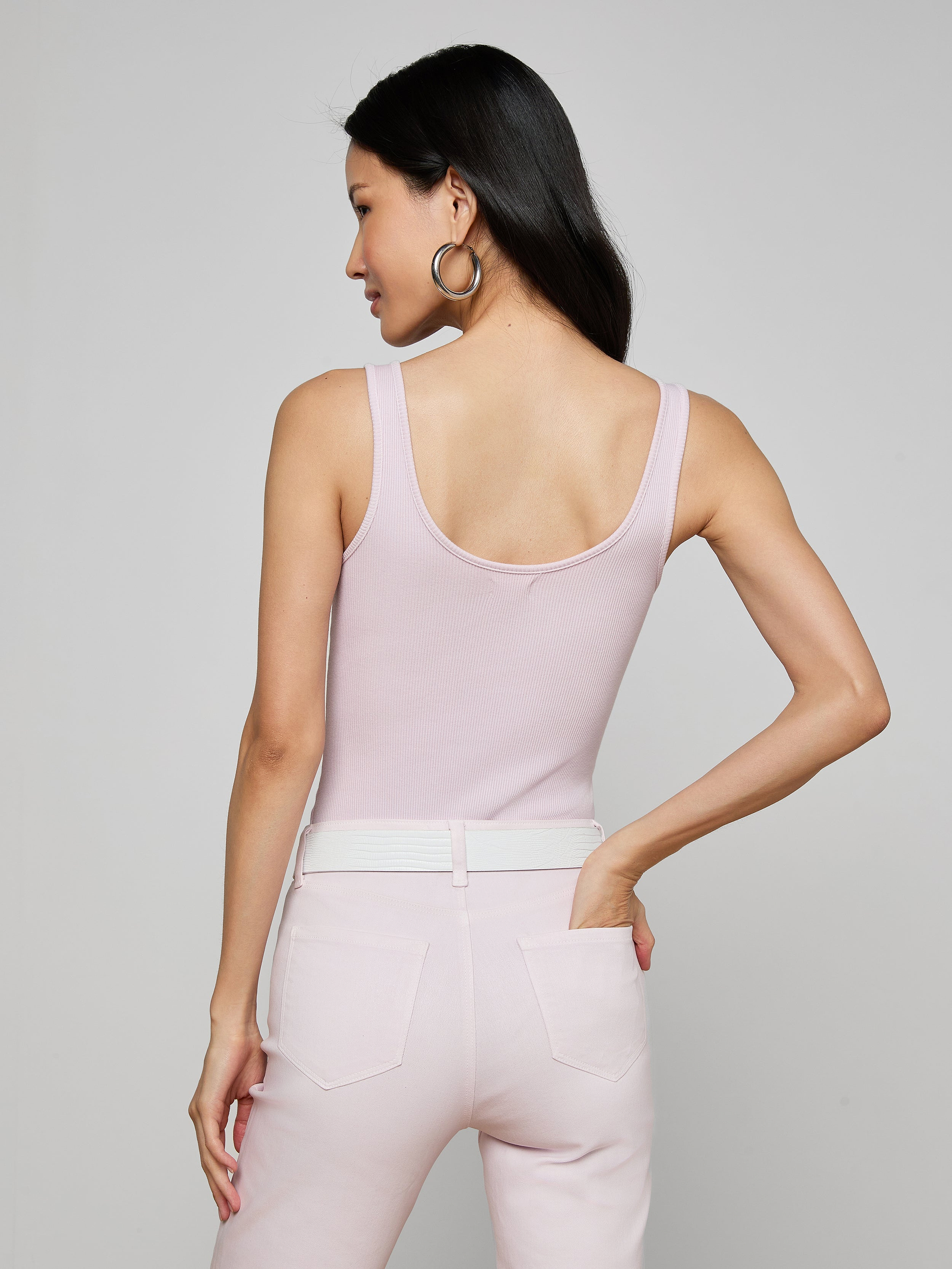 A woman with long dark hair, wearing the Nica Scoopneck Bodysuit in light pink and matching pants, stands with her back to the camera, hand on her hip, against a plain light background.