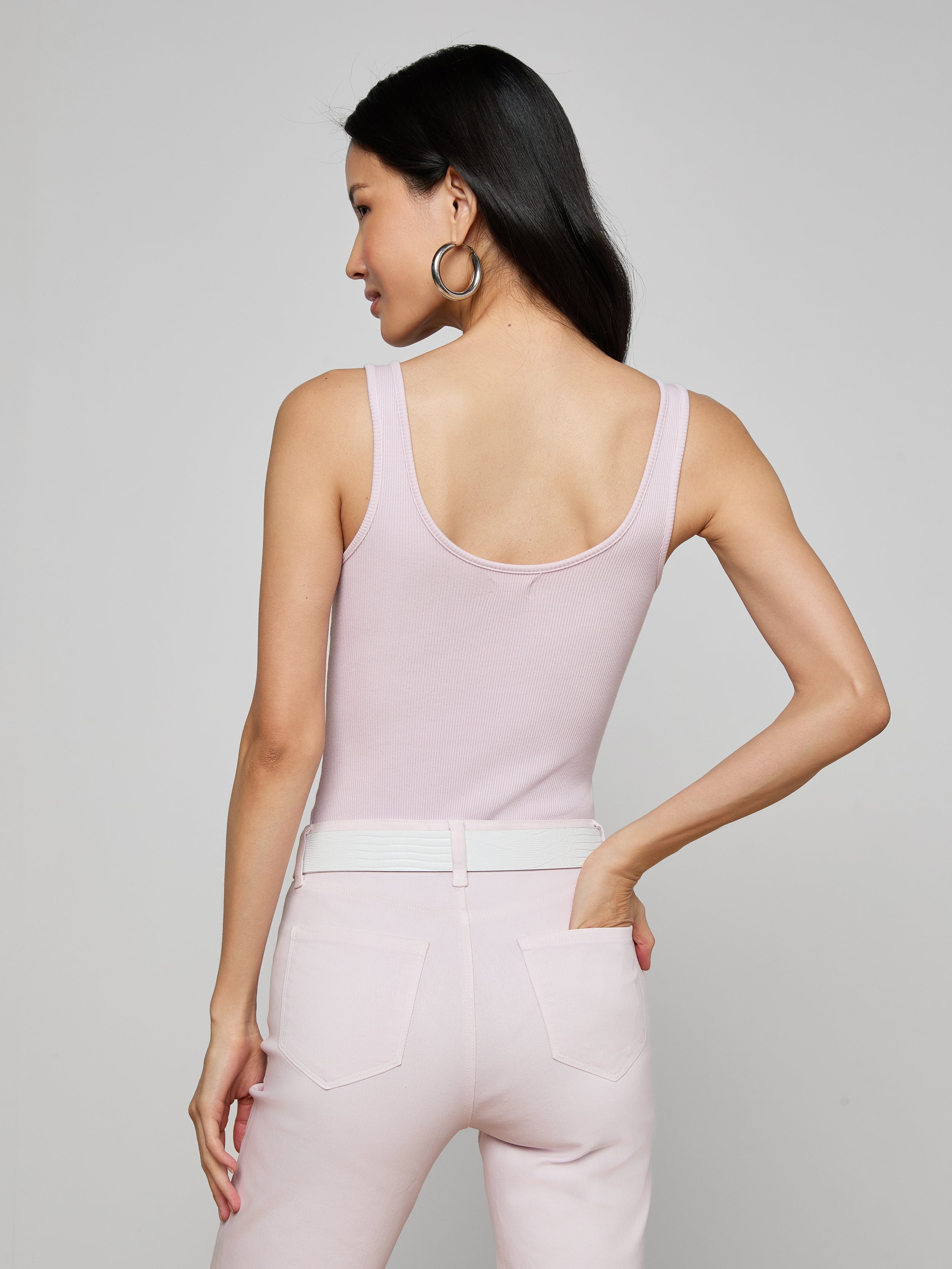 A woman with long dark hair, wearing the Nica Scoopneck Bodysuit in light pink and matching pants, stands with her back to the camera, hand on her hip, against a plain light background.