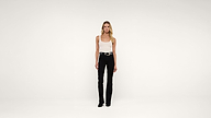 A woman stands facing forward against a plain white background, wearing a white tank top, black pants, black shoes, and a black belt. She has long blonde hair and a neutral expression.