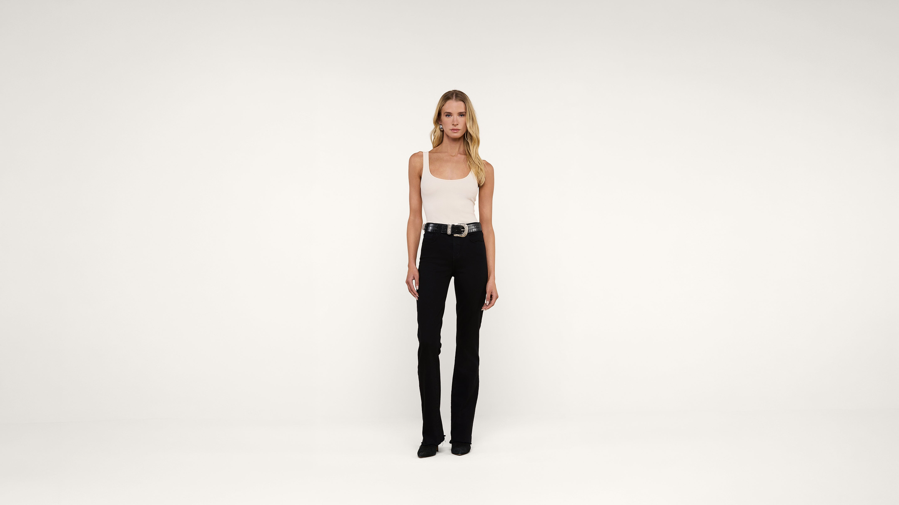 A woman stands facing forward against a plain white background, wearing a white tank top, black pants, black shoes, and a black belt. She has long blonde hair and a neutral expression.