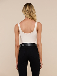 A woman with long blonde hair stands with her back to the camera against a light background, wearing the Nica Scoopneck Bodysuit—a sleeveless, light beige top with a scoop neckline—paired with black pants and a textured black belt. Crafted from stretch-cotton for added comfort, the bodysuit makes an ideal layering piece.