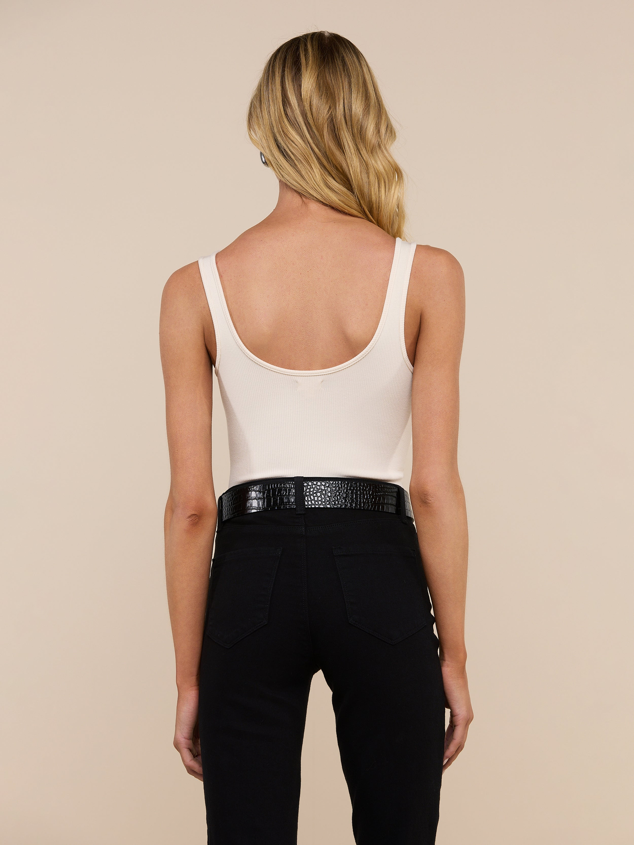 A woman with long blonde hair stands with her back to the camera against a light background, wearing the Nica Scoopneck Bodysuit—a sleeveless, light beige top with a scoop neckline—paired with black pants and a textured black belt. Crafted from stretch-cotton for added comfort, the bodysuit makes an ideal layering piece.
