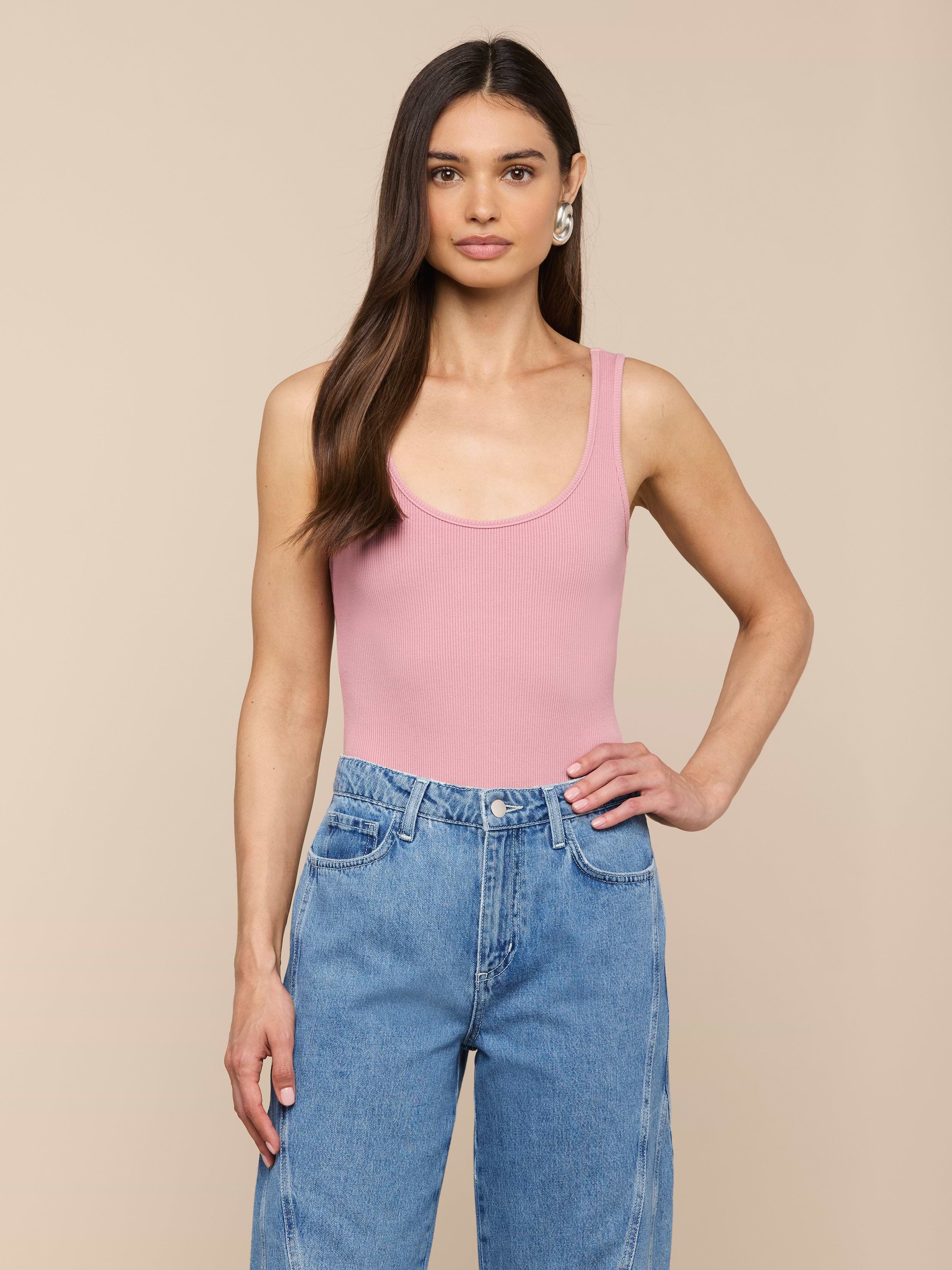 Une femme aux longs cheveux bruns porte un body rose Nica Scoopneck et un jean bleu taille haute, debout sur un fond beige, une main sur la hanche et une expression neutre.