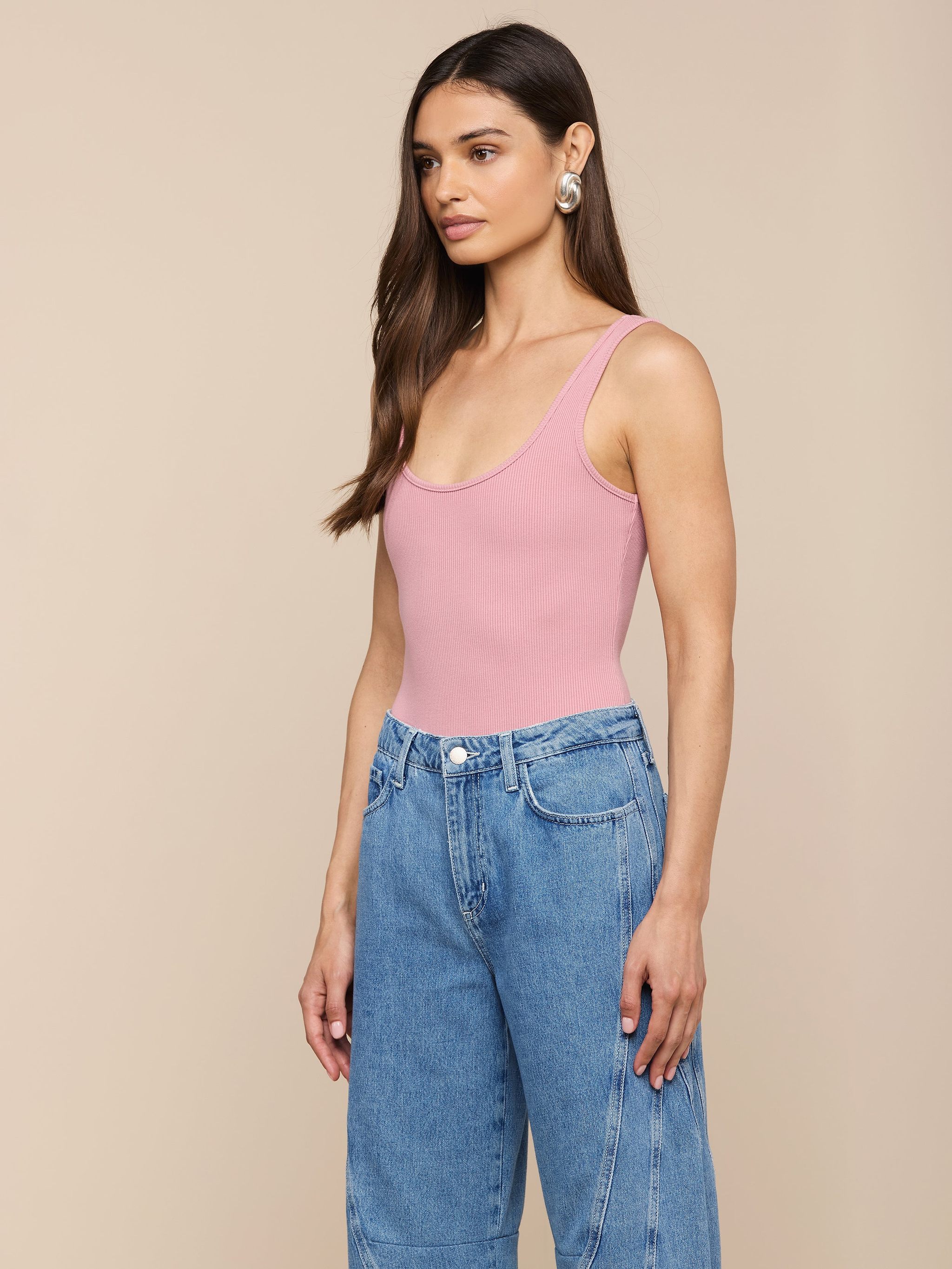 Une femme aux longs cheveux bruns, vêtue dun body rose Nica Scoopneck, dun jean bleu à taille haute et de grandes boucles doreilles argentées, se tient devant un fond beige uni.