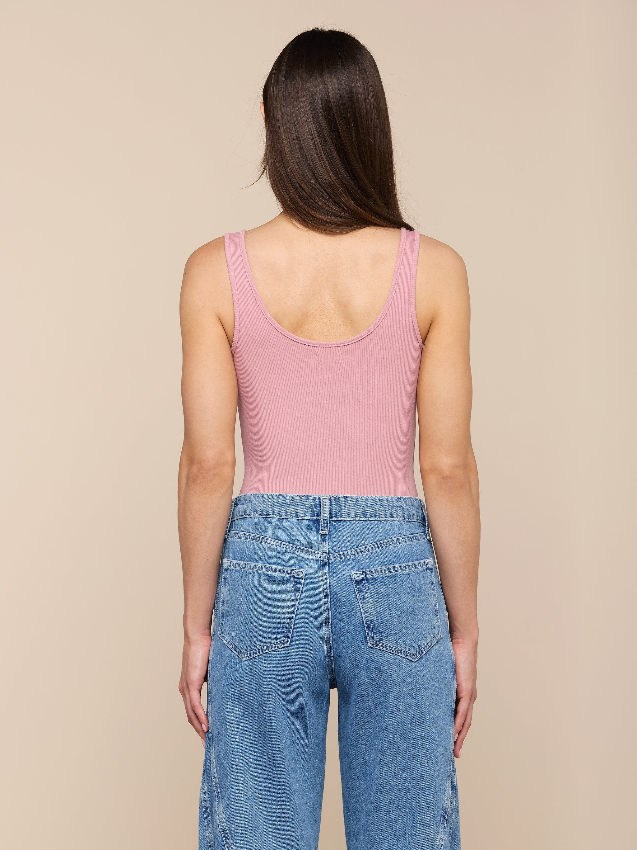 Une personne aux longs cheveux bruns, vêtue dun body rose Nica Scoopneck et dun jean bleu sculpté à taille haute, se tient de face sur un fond beige.