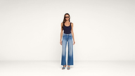 A woman in sunglasses, a navy tank top, and high-waisted, wide-leg jeans stands facing forward against a plain white background. She wears teal open-toe heels.