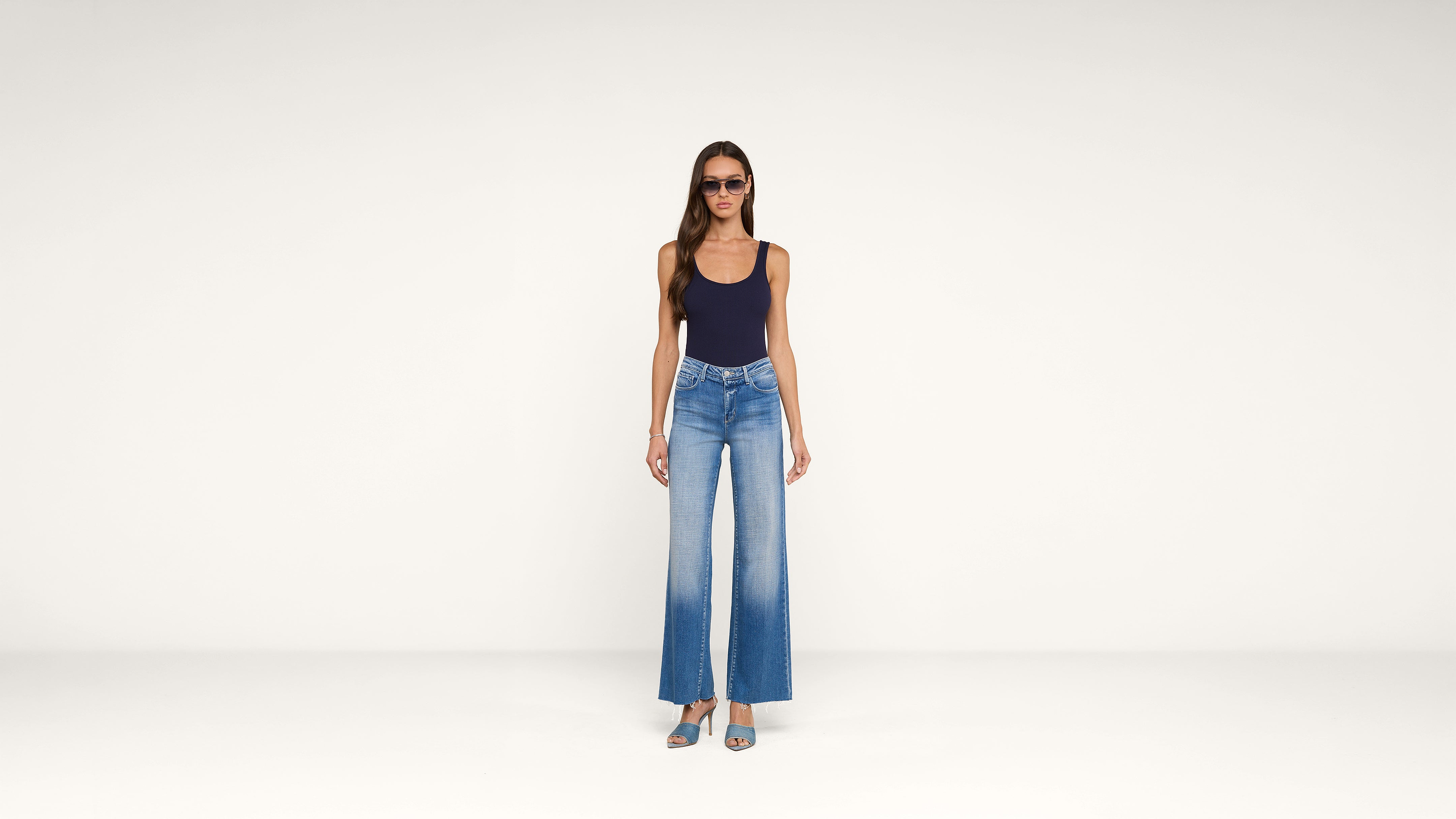 A woman in sunglasses, a navy tank top, and high-waisted, wide-leg jeans stands facing forward against a plain white background. She wears teal open-toe heels.