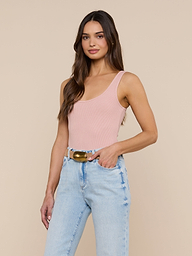 A woman with long brown hair stands and smiles against a plain, off-white background, wearing a light pink Nica Scoopneck Bodysuit with a sculpted fit and light blue high-waisted jeans accessorized with a belt.