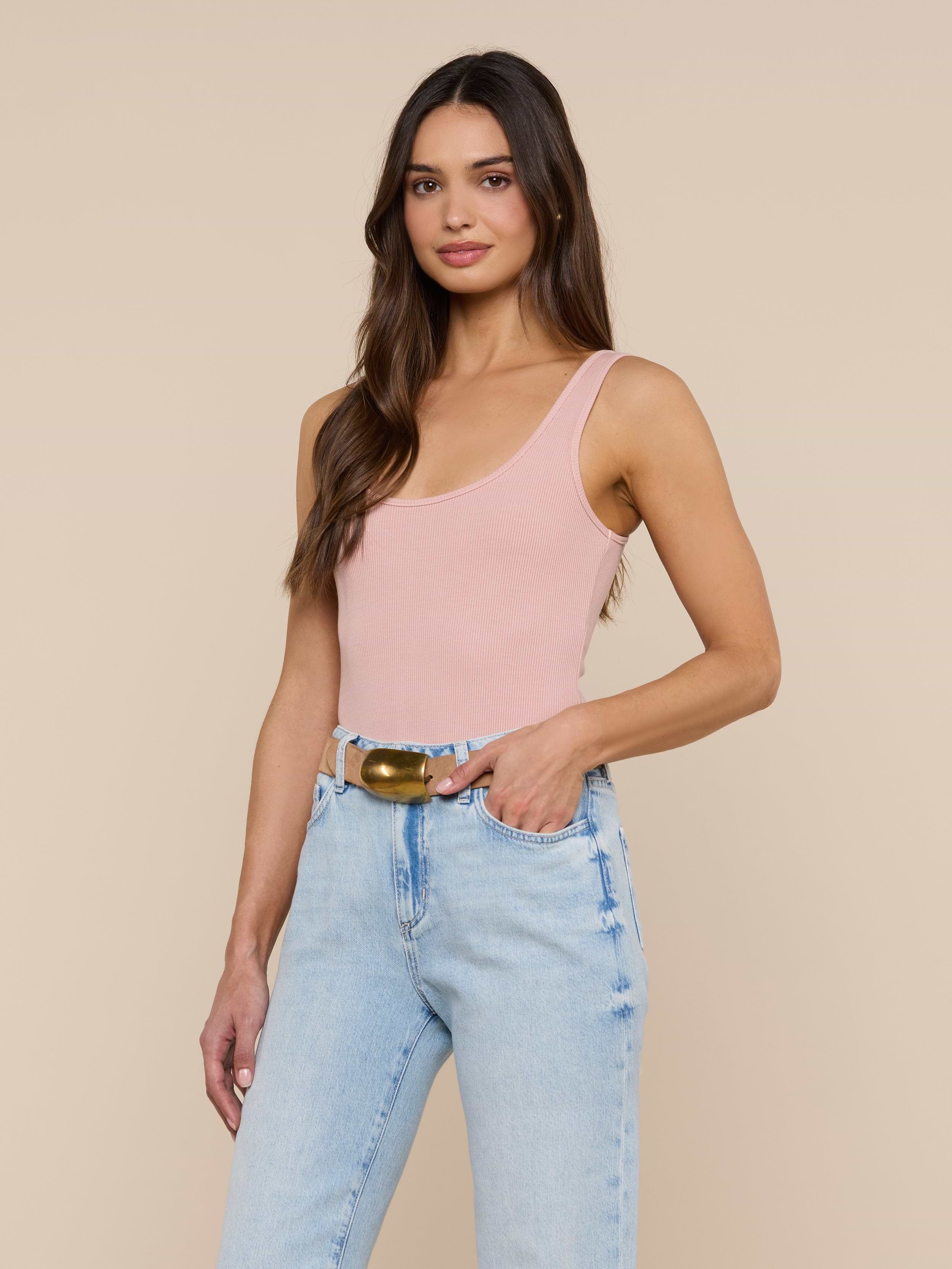 A woman with long brown hair stands and smiles against a plain, off-white background, wearing a light pink Nica Scoopneck Bodysuit with a sculpted fit and light blue high-waisted jeans accessorized with a belt.