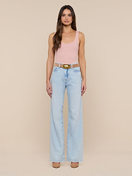 A woman with long brown hair and a neutral expression stands against a plain white background, wearing the Nica Scoopneck Bodysuit in light pink, paired with light blue high-waisted wide-leg jeans, a brown belt, and beige shoes.