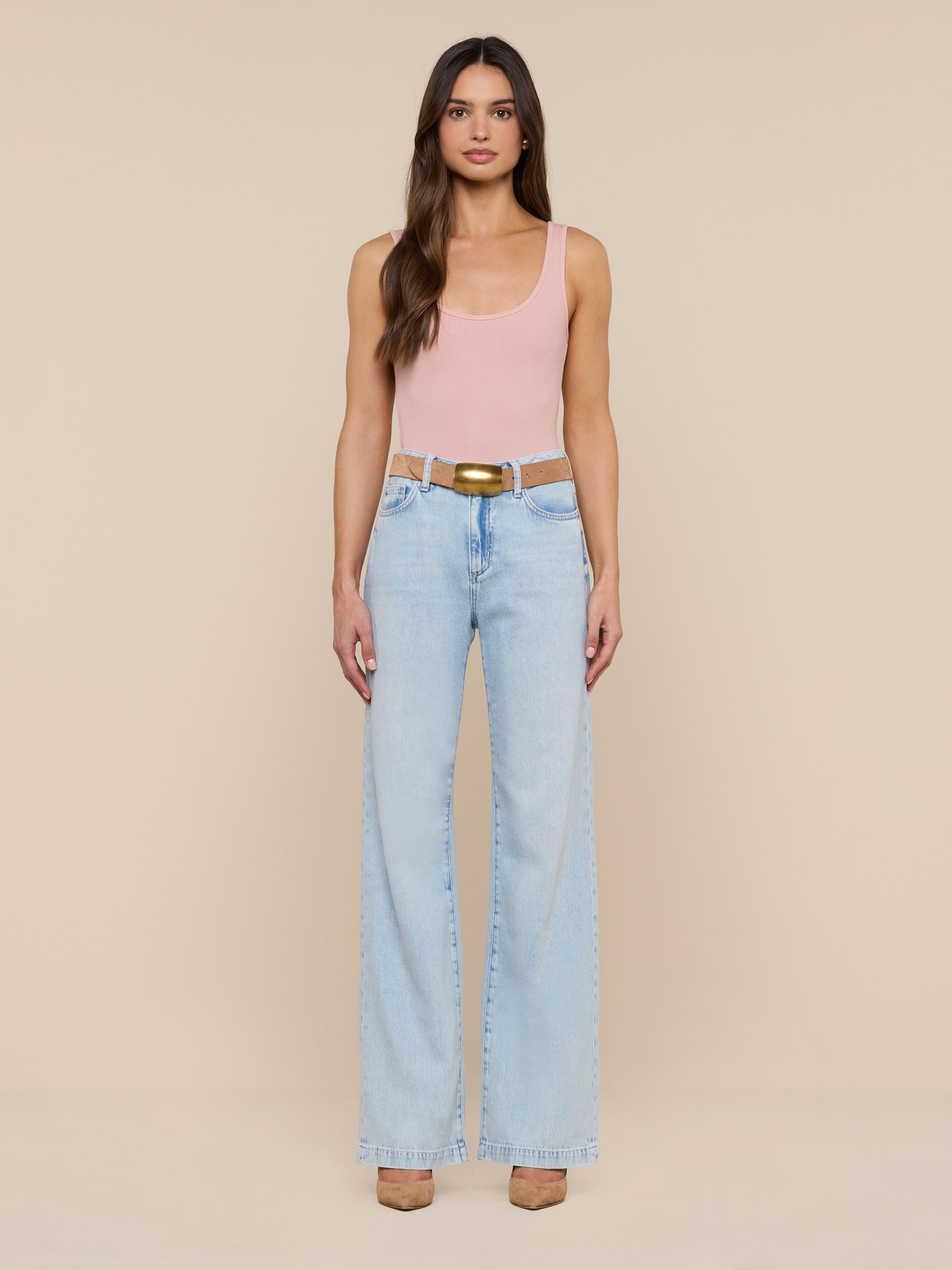 A woman with long brown hair and a neutral expression stands against a plain white background, wearing the Nica Scoopneck Bodysuit in light pink, paired with light blue high-waisted wide-leg jeans, a brown belt, and beige shoes.