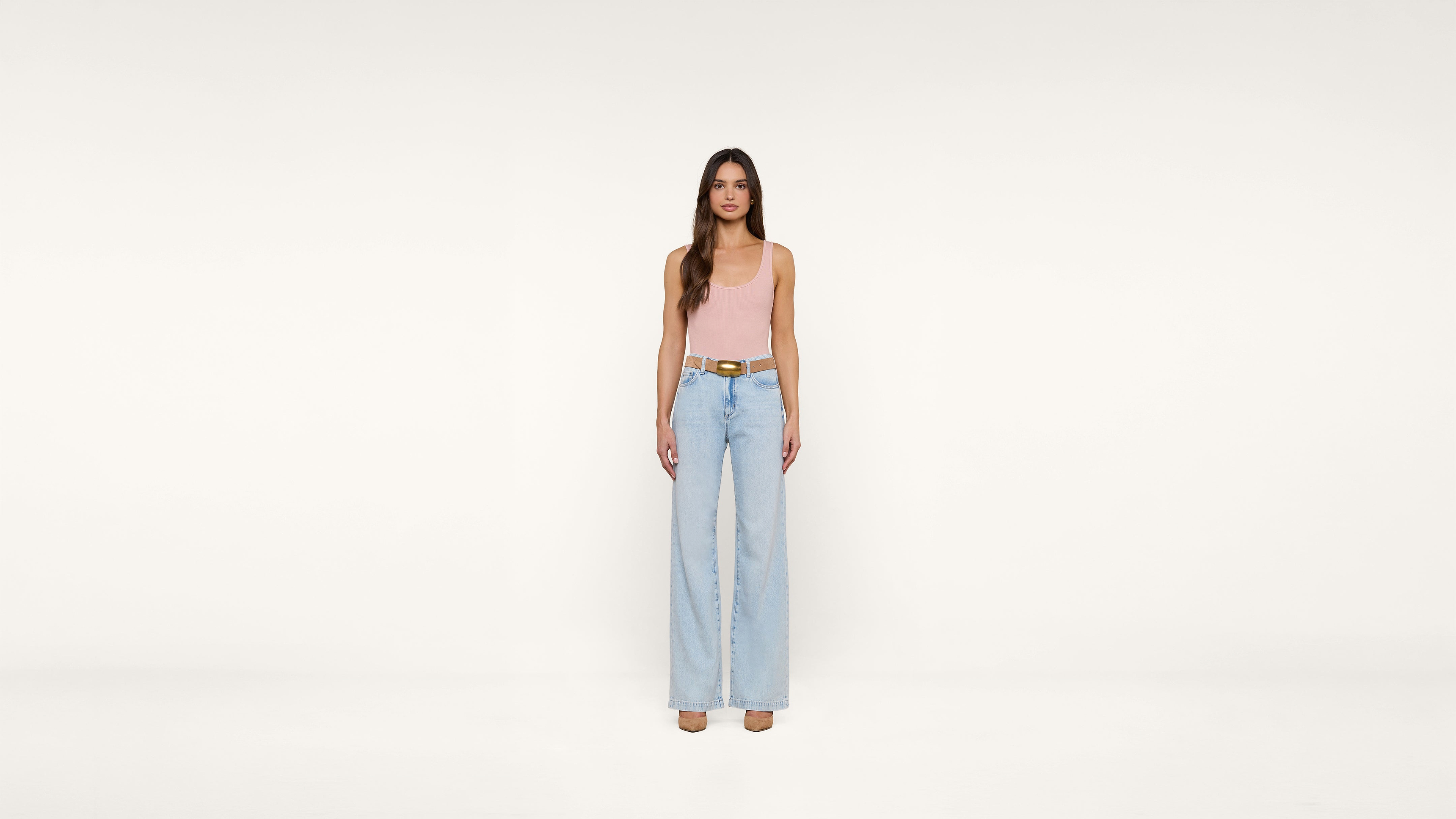 A woman stands against a plain white background, wearing a pink sleeveless top, light blue wide-leg jeans, a brown belt, and beige shoes. She has long brown hair and a neutral expression.