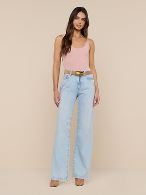 A woman stands against a beige background, wearing a pink Nica Scoopneck Bodysuit with a sculpted fit, light blue high-waisted wide-leg jeans, a brown belt, and tan shoes. She has long wavy brown hair and a relaxed expression.