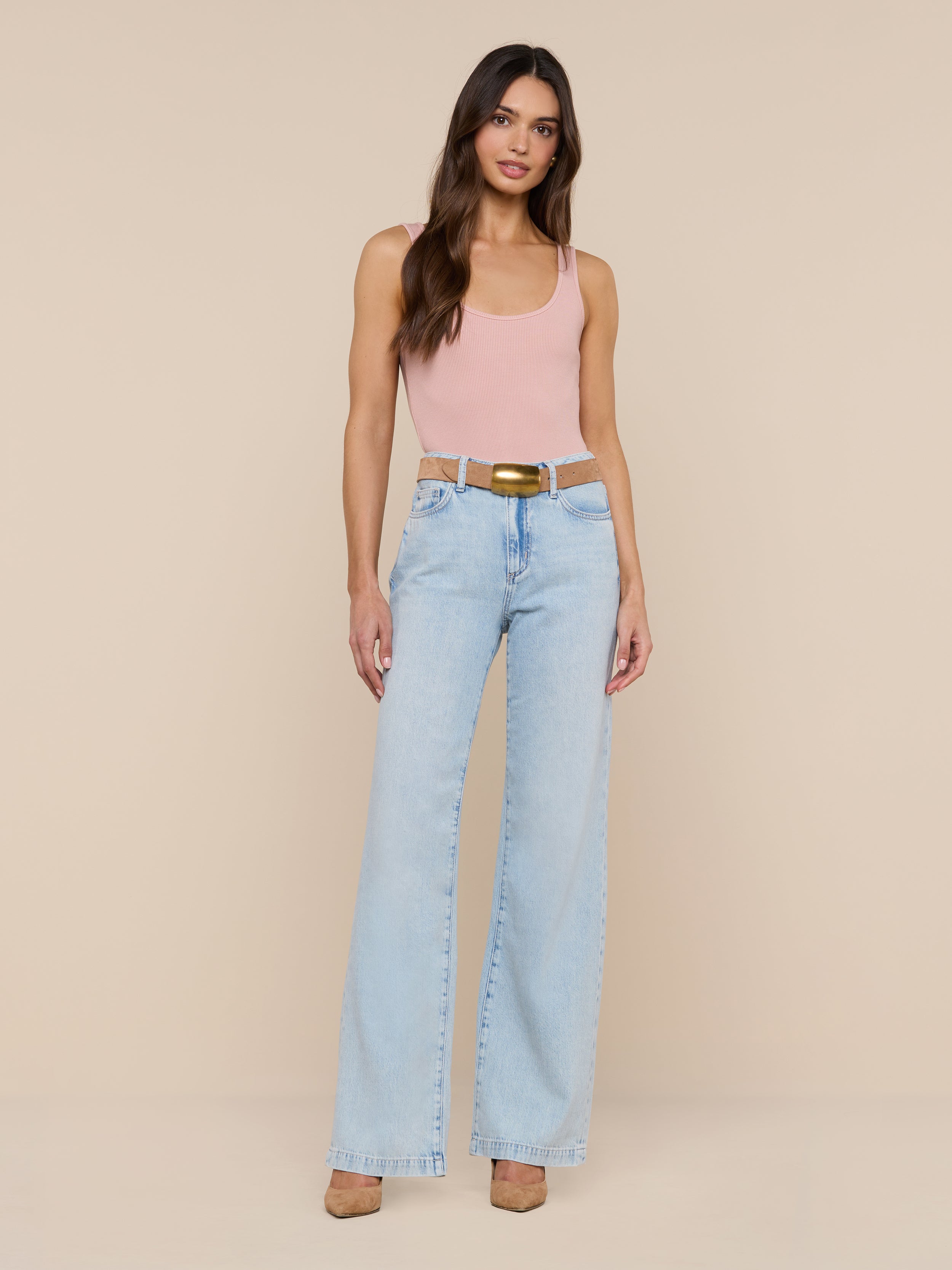 A woman stands against a plain white background, wearing a light pink Nica Scoopneck Bodysuit with a sculpted fit, light blue high-waisted wide-leg jeans, and beige high heels.
