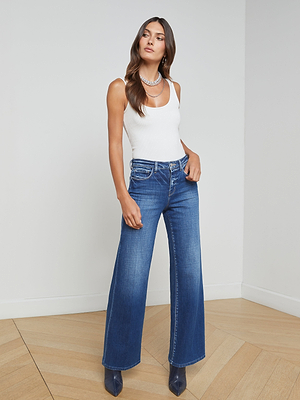 A woman with long brown hair poses indoors in the Nica Scoopneck Bodysuit, blue wide-leg jeans, pointed-toe shoes, and layered necklaces. She stands with one hand in her pocket against a light wall and wood floor.