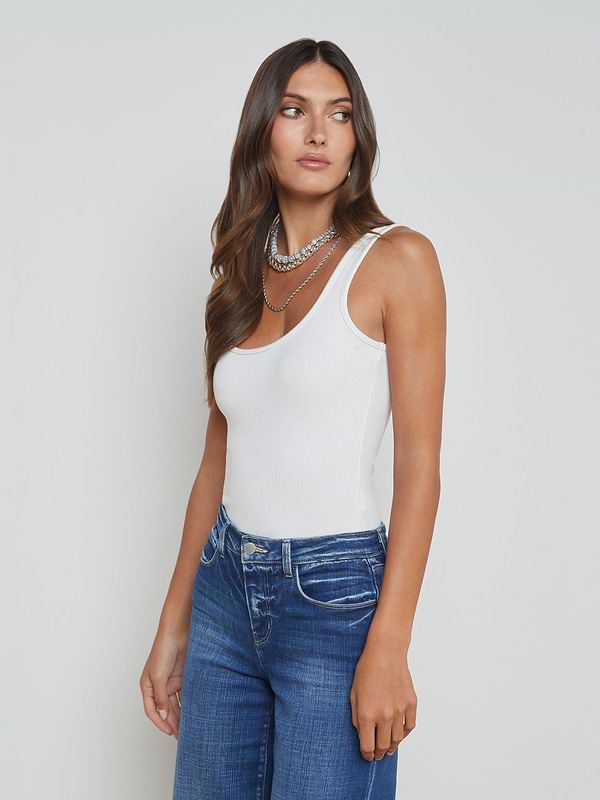 A woman with long brown hair wears the Nica Scoopneck Bodysuit and blue jeans, accessorized with layered silver necklaces. She stands against a plain white background, facing slightly to the side and looking off to her left.