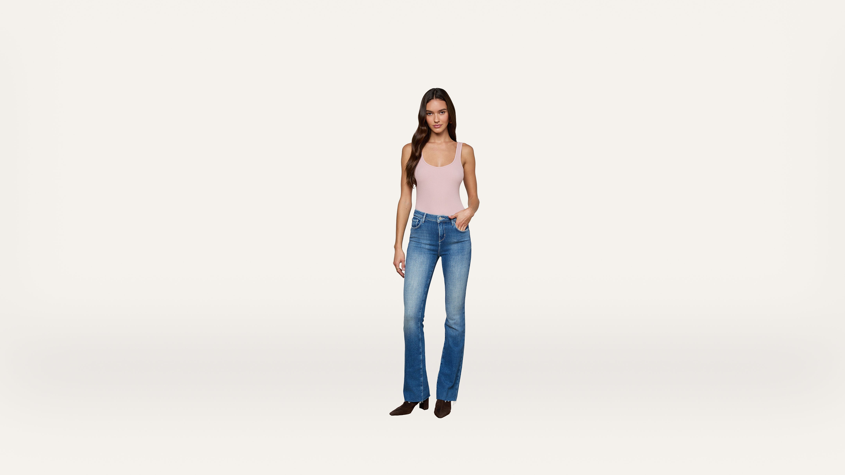 A woman wearing a light pink tank top, blue jeans, and black shoes stands against a plain white background, with one hand in her pocket.