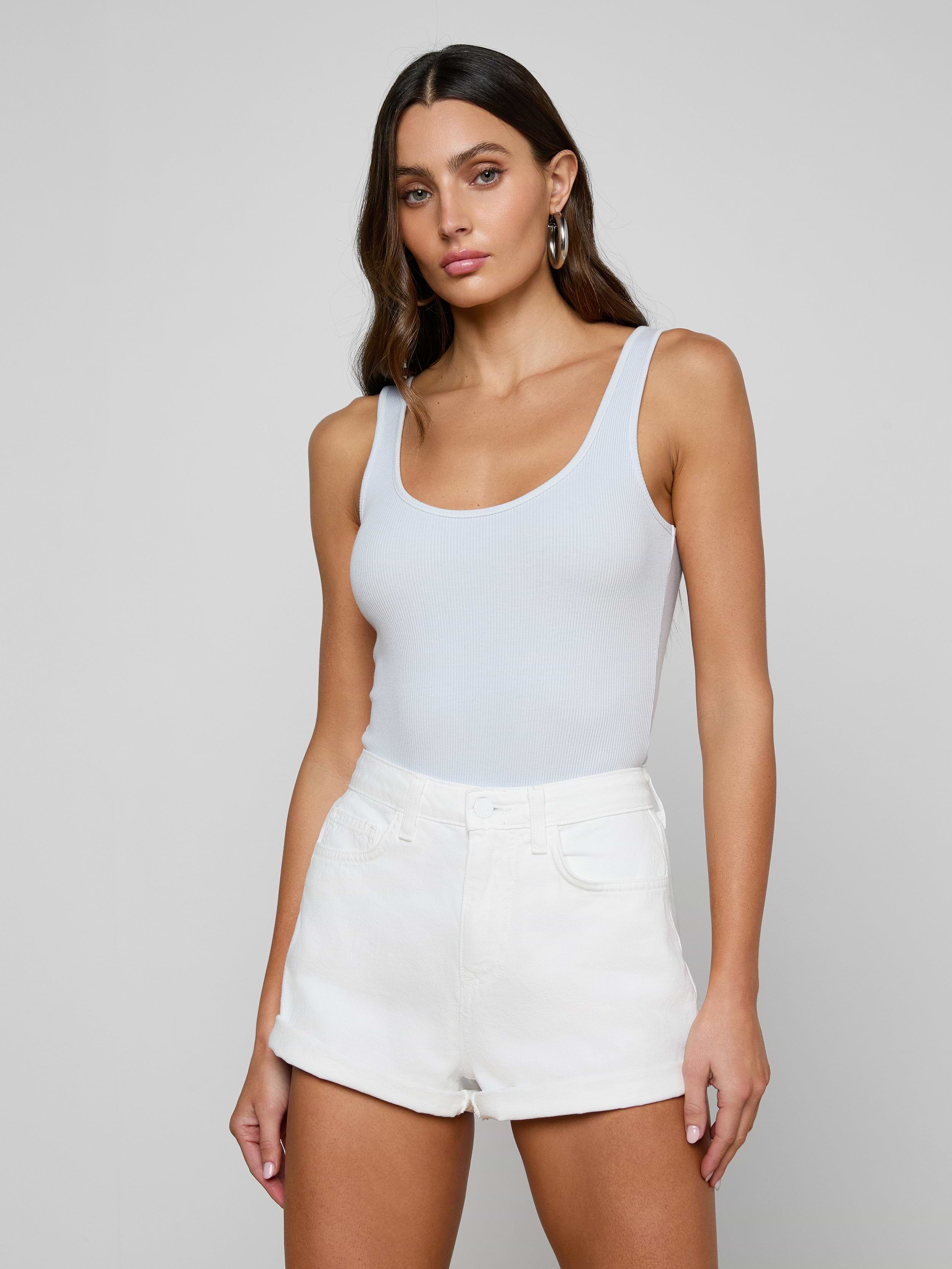 A woman with long brown hair wears a white Nica Scoopneck Bodysuit and high-waisted white shorts, paired with gold hoop earrings, standing against a plain light gray background with a neutral expression.