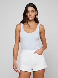 A woman with long brown hair wears a white Nica Scoopneck Bodysuit and white shorts, standing against a light gray background with one hand in her pocket, highlighting the sculpted fit of the high-recovery stretch rib fabric.