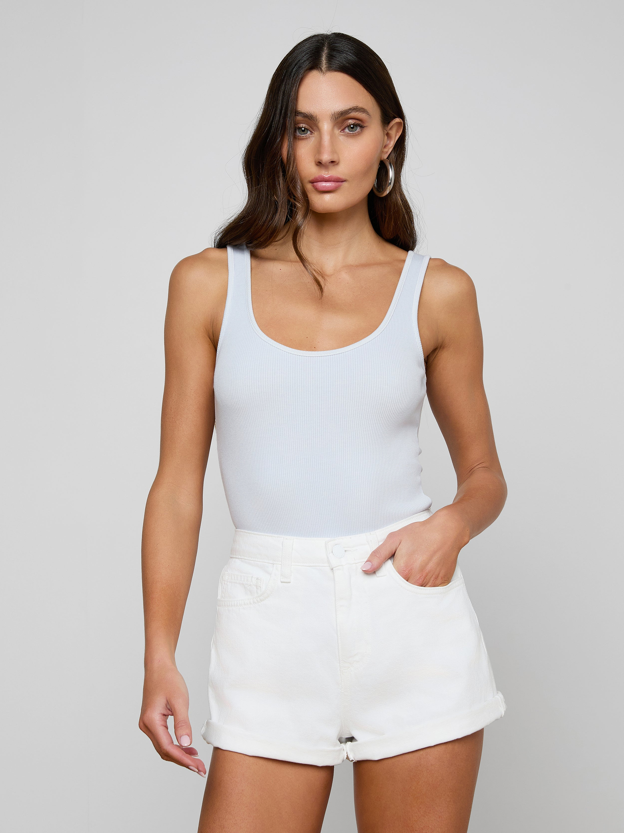 A woman with long brown hair wears a white Nica Scoopneck Bodysuit and white shorts, standing against a light gray background with one hand in her pocket, highlighting the sculpted fit of the high-recovery stretch rib fabric.
