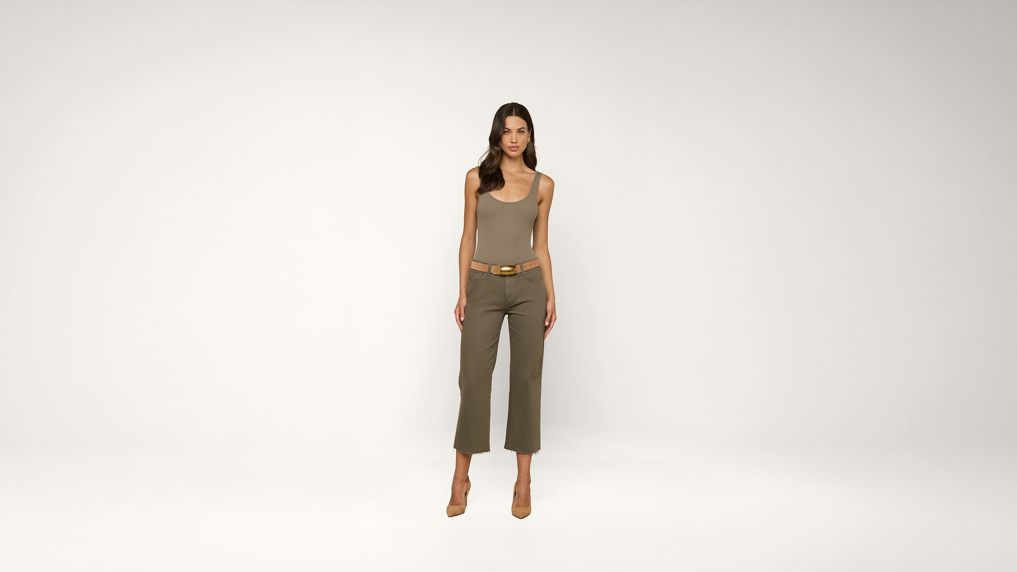 A woman stands against a plain white background, wearing a sleeveless ribbed top, belted olive green pants, and brown heels.