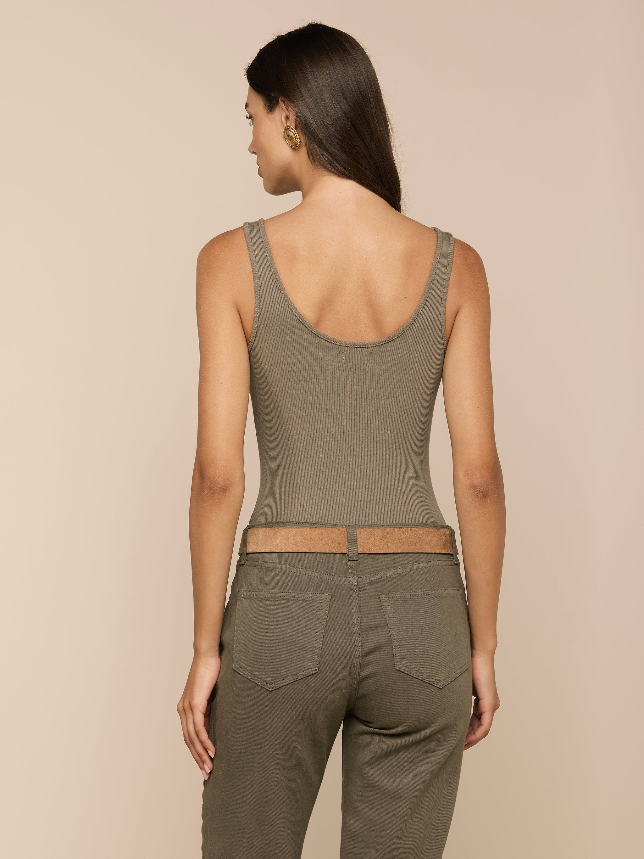 A woman stands facing away, wearing the Nica Scoopneck Bodysuit in olive—a sleeveless, sculpted-fit top with a scoop neckline—paired with matching high-waisted pants. Her long hair is loose, and she accessorizes with gold hoop earrings against a plain, light background.