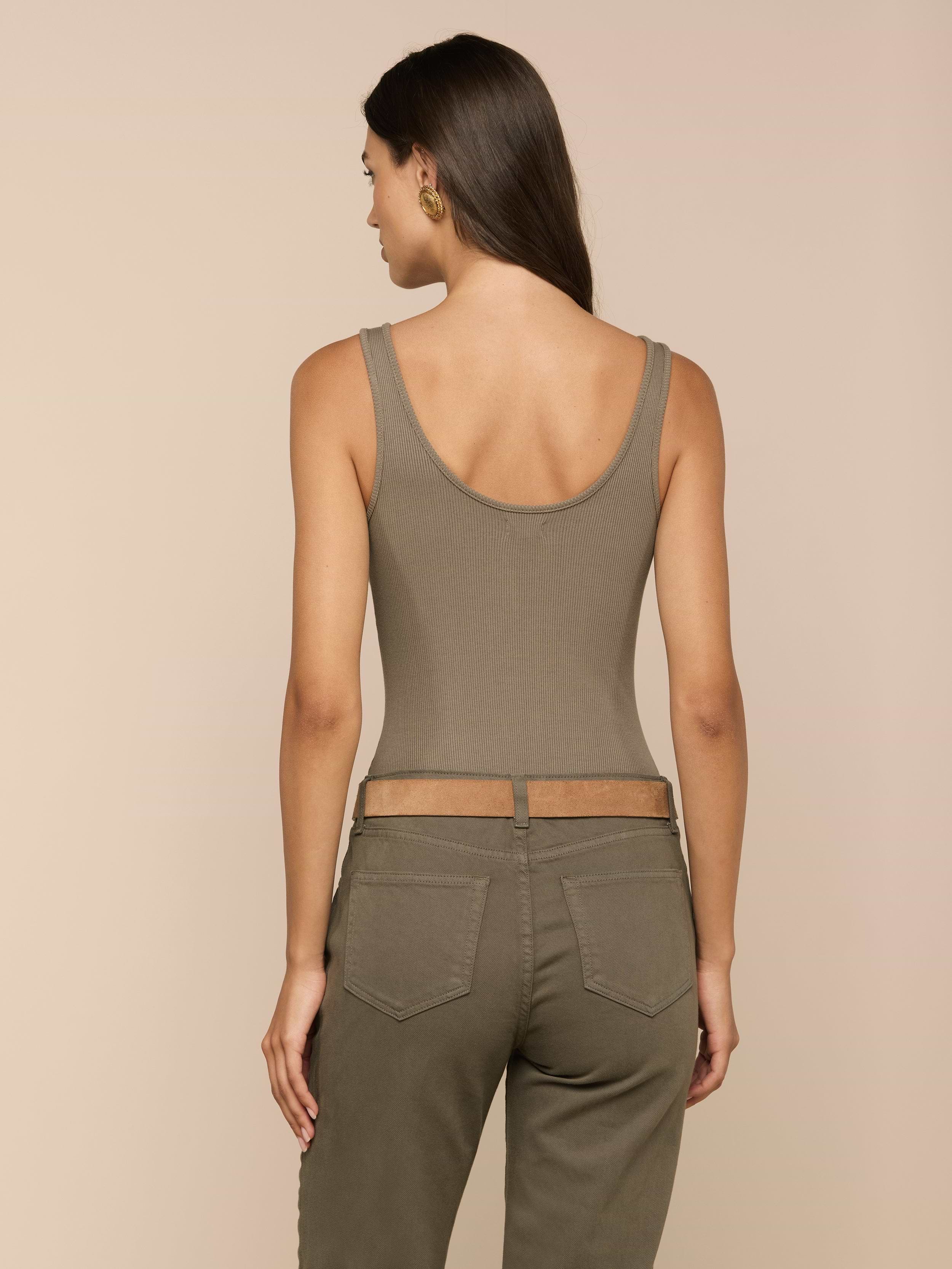 A woman stands facing away, wearing the Nica Scoopneck Bodysuit in olive—a sleeveless, sculpted-fit top with a scoop neckline—paired with matching high-waisted pants. Her long hair is loose, and she accessorizes with gold hoop earrings against a plain, light background.