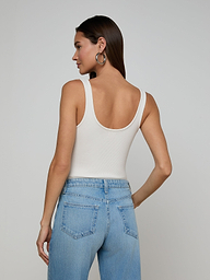 A woman with long blonde hair stands facing away, wearing a sleeveless beige Nica Scoopneck Bodysuit tucked into high-waisted black pants with a black textured belt, set against a plain light background.