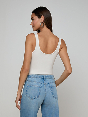 A woman with long brown hair and large hoop earrings wears the Nica Scoopneck Bodysuit and high-waisted light blue jeans, standing with her back to the camera against a plain light background.