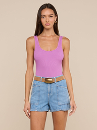 A woman stands against a plain light background wearing a sleeveless pink Nica Scoopneck Bodysuit made of high recovery stretch rib, paired with light blue denim shorts featuring large front pockets and a brown belt.