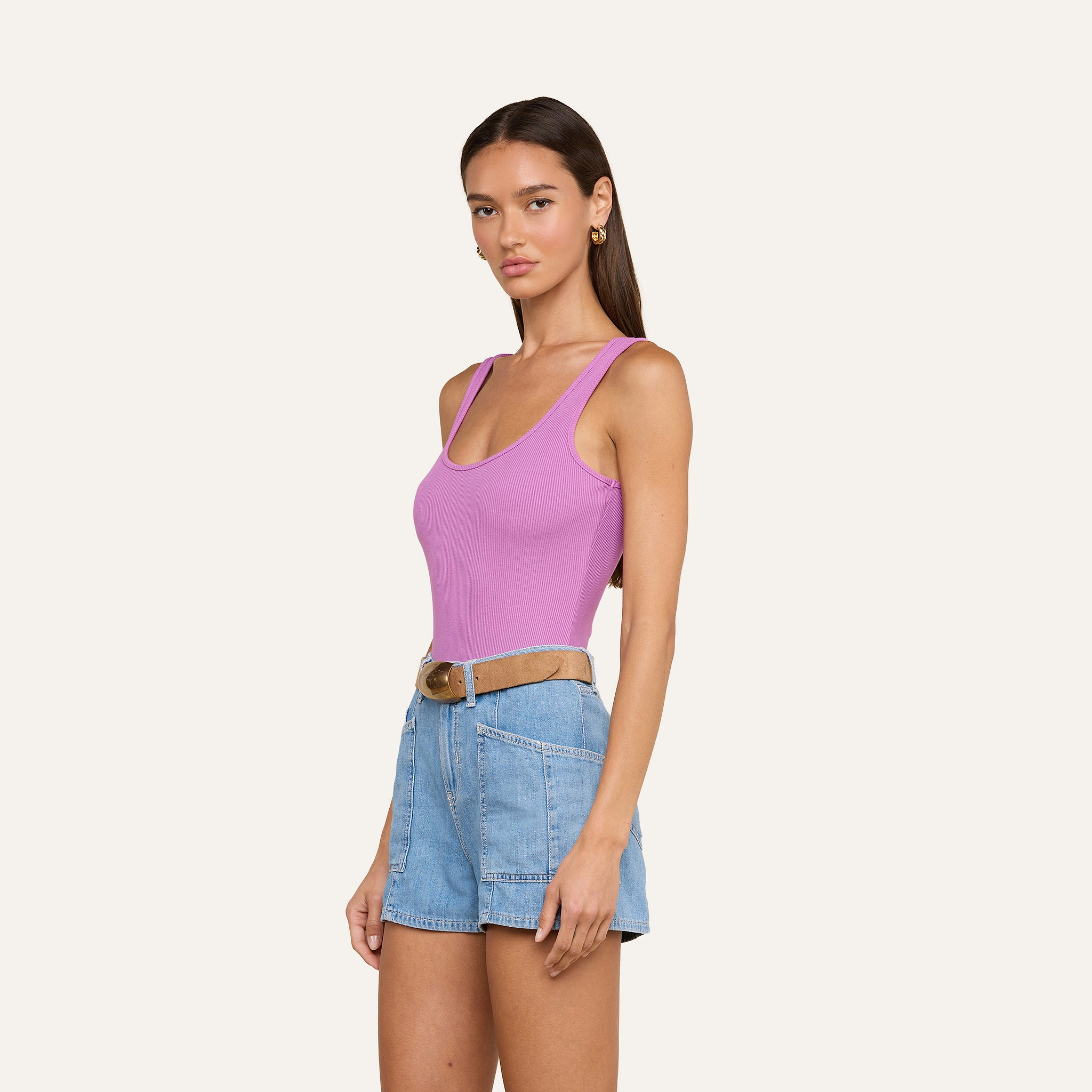 A woman stands against a plain light background wearing the Nica Scoopneck Bodysuit in pink, featuring a sleeveless design and crafted from high recovery stretch rib fabric that sculpts her silhouette. She pairs it with high-waisted denim shorts and a brown belt.