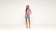 A woman stands against a plain white background, wearing a pink tank top, denim shorts, a belt, and nude heels. One hand is in her pocket and she faces the camera.