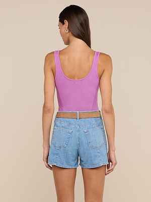 A woman with long brown hair and gold hoop earrings faces away, wearing a pink Nica Scoopneck Bodysuit and light blue denim shorts against a plain beige background.