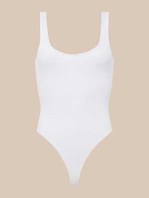 The Nica Scoopneck Bodysuit is a plain white, sleeveless ribbed knit piece featuring a scoop neckline and high-cut legs, shown on a beige background.