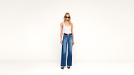 A woman wearing sunglasses, a white tank top, blue wide-leg jeans, and black heels stands against a plain white background.