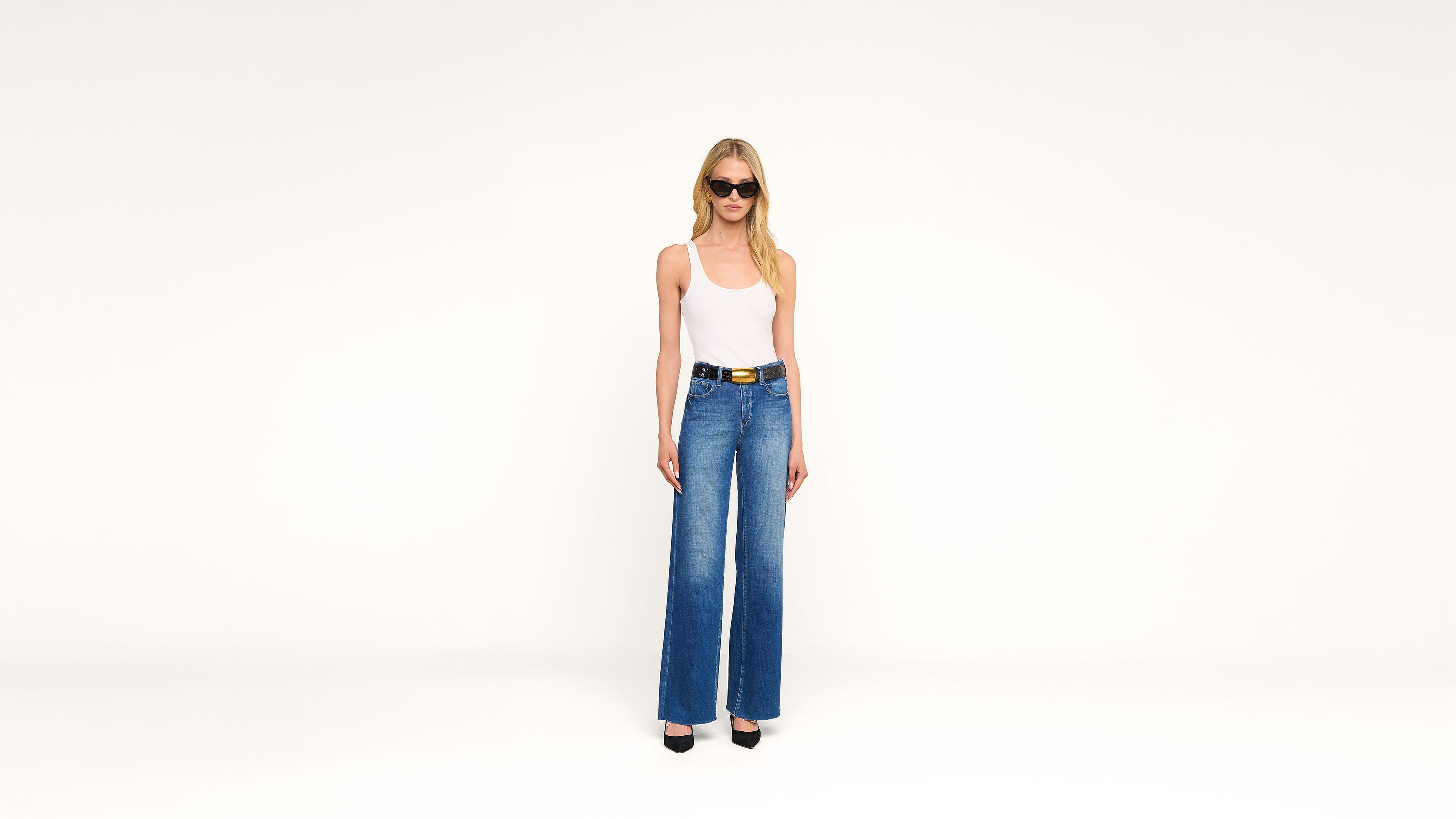 A woman wearing sunglasses, a white tank top, blue wide-leg jeans, and black heels stands against a plain white background.