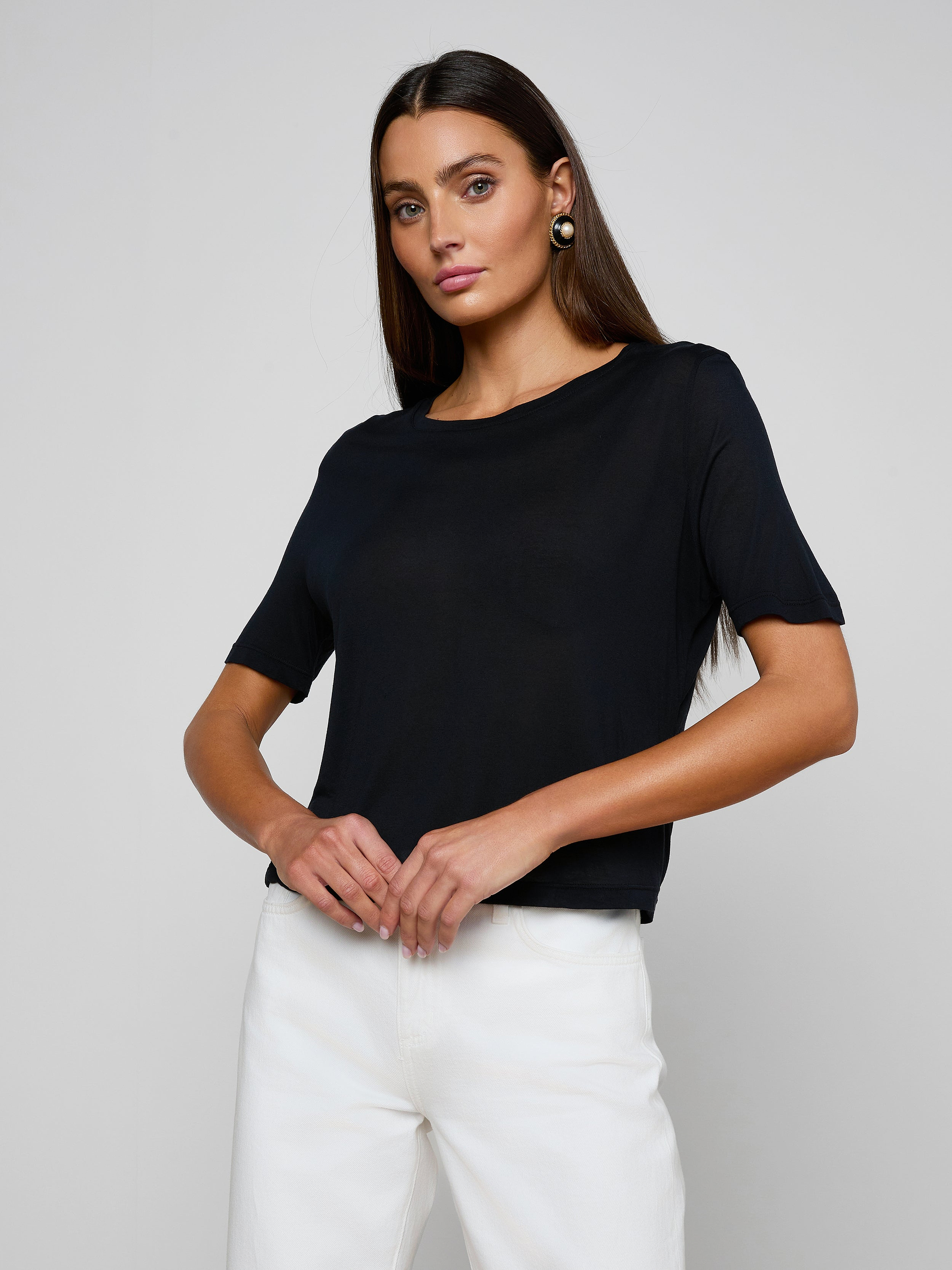 A woman with long dark hair stands confidently against a plain light background, wearing the Remy Tee—a relaxed fit, black short-sleeve top in micro modal jersey—and white pants, her hands resting at her waist as she looks at the camera.