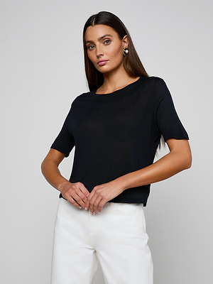 A woman with long brown hair models the Remy Tee in black and white pants, standing against a light gray background with her hands loosely clasped, gazing slightly to the side.