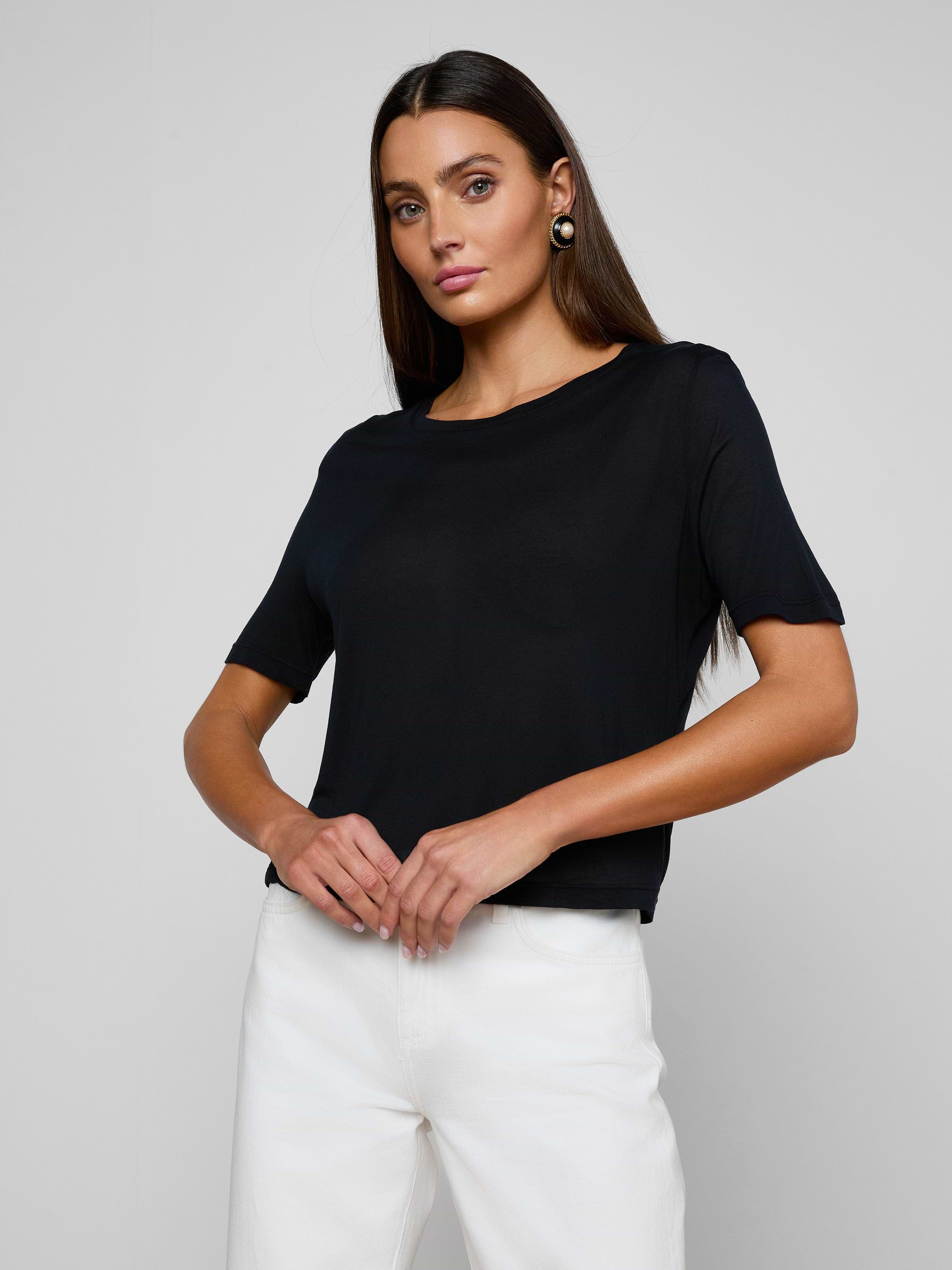 A woman with long dark hair stands confidently against a plain light background, wearing the Remy Tee—a relaxed fit, black short-sleeve top in micro modal jersey—and white pants, her hands resting at her waist as she looks at the camera.